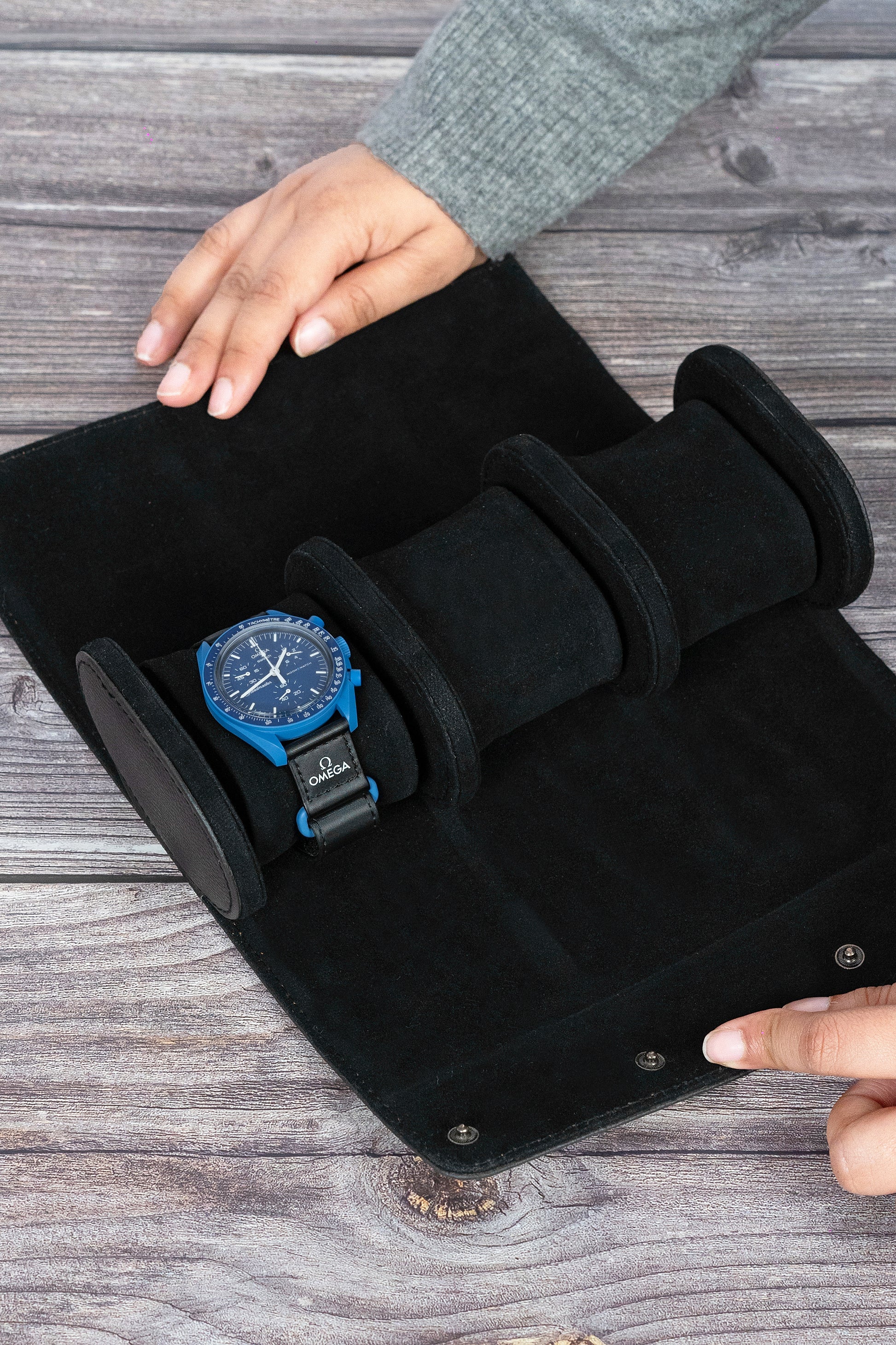 A black vegan leather watch box with a blue watch resting on the cushioned interior, being held open by a person.